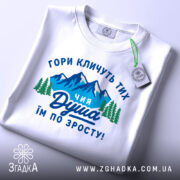 Футболка душа гір. Біла футболка, складена на поверхні, з принтом, що зображає гори та дерева, текст на українській мові: 'ГОРИ КЛИЧУТЬ ТИХ ЧИЯ ДУША ІМ ПО ЗРОСТУ!'. На футболці є етикетка з зеленим шнурком, фон світло-сірий.