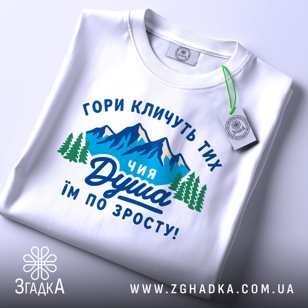 Футболка душа гір. Біла футболка, складена на поверхні, з принтом, що зображає гори та дерева, текст на українській мові: 'ГОРИ КЛИЧУТЬ ТИХ ЧИЯ ДУША ІМ ПО ЗРОСТУ!'. На футболці є етикетка з зеленим шнурком, фон світло-сірий.