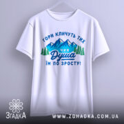 Футболка душа гір. Біла футболка, підвішена на вішалці, з принтом, що зображає гори та дерева, текст на українській мові: 'ГОРИ КЛИЧУТЬ ТИХ ЧИЯ ДУША ІМ ПО ЗРОСТУ!'. Фон світло-сірий, футболка має короткі рукави.