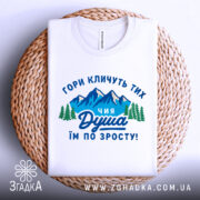 Футболка душа гір. Біла футболка, складена на плетеній основі, з принтом, що зображає гори та дерева, текст на українській мові: 'ГОРИ КЛИЧУТЬ ТИХ ЧИЯ ДУША ІМ ПО ЗРОСТУ!'. Фон світло-сірий.