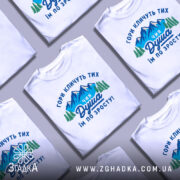 Футболка душа гір. Біла футболка на манекені, з принтом, що зображає гори та дерева, текст на українській мові: 'ГОРИ КЛИЧУТЬ ТИХ ЧИЯ ДУША ІМ ПО ЗРОСТУ!'. Фон світло-сірий, футболка має короткі рукави.