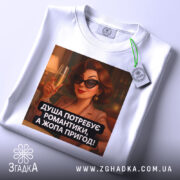 Футболка душа потребує романтики. Біла футболка, зображена зверху, принт з жінкою в чорних окулярах, яка тримає келих з шампанським. На футболці є етикетка з логотипом бренду. Текст принта: 'ДУША ПОТРЕБУЄ РОМАНТИКИ, А ЖОПА ПРИГОД!'