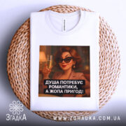 Футболка душа потребує романтики. Біла футболка, зображена зверху на плетеній основі, принт з жінкою в чорних окулярах, яка тримає келих з шампанським. Текст принта: 'ДУША ПОТРЕБУЄ РОМАНТИКИ, А ЖОПА ПРИГОД!'