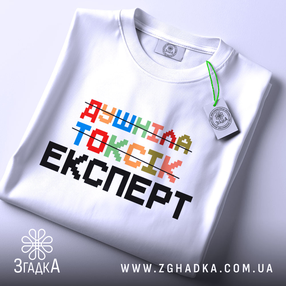 Футболка душніла токсік експерт, білого кольору, зображена зверху, на столі, фон світло-сірий, принт кольоровий з написом 'Душніла токсік експерт' та етикеткою з зеленим шнурком.