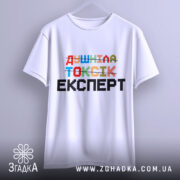 Футболка душніла токсік експерт, білого кольору, зображена з переднього ракурсу на вішалці, фон світло-сірий, принт кольоровий з написом 'Душніла токсік експерт' у різних шрифтах.