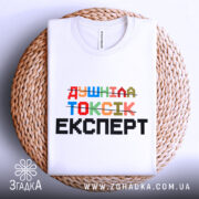 Футболка душніла токсік експерт, білого кольору, зображена зверху на плетеній кошику, фон світло-сірий, принт кольоровий з написом 'Душніла токсік експерт' у різних шрифтах.