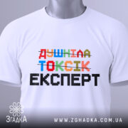 Футболка душніла токсік експерт, білого кольору, зображена зверху, на столі, фон світло-сірий, принт кольоровий з написом 'Душніла токсік експерт' та етикеткою з зеленим шнурком.