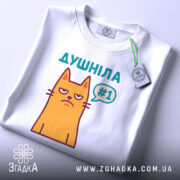 Футболка душніла жарт, біла, складена на поверхні, з верхнього ракурсу, фон світло-сірий, принт з жовтим котом з незадоволеним виразом обличчя та написом "Душніла #1" у кольорах бірюзовий та фіолетовий, з етикеткою на зеленому шнурку.
