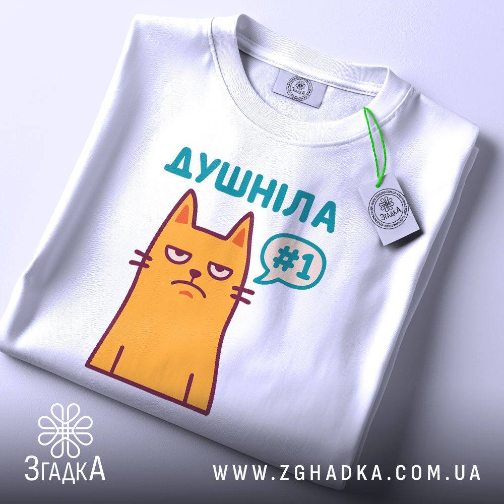 Футболка душніла жарт, біла, складена на поверхні, з верхнього ракурсу, фон світло-сірий, принт з жовтим котом з незадоволеним виразом обличчя та написом "Душніла #1" у кольорах бірюзовий та фіолетовий, з етикеткою на зеленому шнурку.