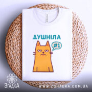 Футболка душніла жарт, біла, складена на плетеній кошику, з верхнього ракурсу, фон світло-сірий, принт з жовтим котом з незадоволеним виразом обличчя та написом "Душніла #1" у кольорах бірюзовий та фіолетовий.