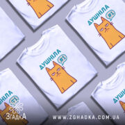 Футболка душніла жарт, біла, зображена на манекені з ближнього ракурсу, фон світло-сірий, принт з жовтим котом з незадоволеним виразом обличчя та написом "Душніла #1" у кольорах бірюзовий та фіолетовий.