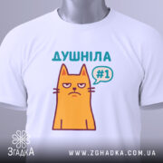 Футболка душніла жарт, біла, складена на поверхні, з верхнього ракурсу, фон світло-сірий, принт з жовтим котом з незадоволеним виразом обличчя та написом "Душніла #1" у кольорах бірюзовий та фіолетовий, з етикеткою на зеленому шнурку.