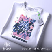 Футболка дикий стиль, біла, зображена зверху, фон світло-сірий, принт з квітами та написом 'Wild Style', на футболці є етикетка з зеленим шнурком.