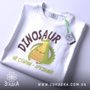 Футболка динозавр не старий. Білий колір, зображена зверху, складена на поверхні. Фон світло-сірий, принт з динозавром та написом 'НЕ СТАРИЙ - АРХІВНИЙ!' у коричневих тонах, з етикеткою на шнурку.