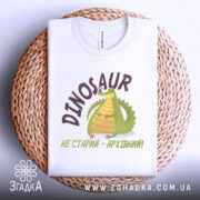 Футболка динозавр не старий. Білий колір, зображена зверху на плетеній основі. Фон білий, принт з динозавром та написом 'НЕ СТАРИЙ - АРХІВНИЙ!' у коричневих тонах.