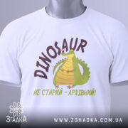Футболка динозавр не старий. Білий колір, зображена зверху, складена на поверхні. Фон світло-сірий, принт з динозавром та написом 'НЕ СТАРИЙ - АРХІВНИЙ!' у коричневих тонах, з етикеткою на шнурку.