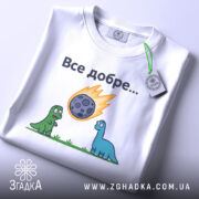 Футболка динозаври все добре. Біла футболка, складена на столі, зображена з верхнього ракурсу. На фоні світло-сірий градієнт. Принт з двома динозаврами, один зелений, інший блакитний, які дивляться на метеорит, що падає, з написом 'Все добре...' над ними. На футболці є етикетка.