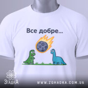 Футболка динозаври все добре. Біла футболка, складена на столі, зображена з верхнього ракурсу. На фоні світло-сірий градієнт. Принт з двома динозаврами, один зелений, інший блакитний, які дивляться на метеорит, що падає, з написом 'Все добре...' над ними. На футболці є етикетка.