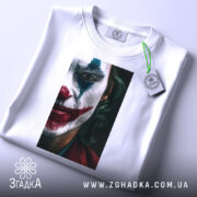 Футболка Джокер білого кольору, складена на поверхні, фон світло-сірий. Принт з обличчям Джокера, видно деталі макіяжу, з етикеткою, що звисає з коміра.