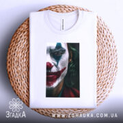 Футболка Джокер білого кольору, складена на плетеній кошику, фон світло-сірий. Принт з обличчям Джокера, видно деталі макіяжу, тканина футболки гладка.