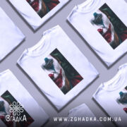 Футболка Джокер білого кольору, зображена на манекені з блискучою поверхнею, фон світло-сірий. Принт з обличчям Джокера з червоними губами та зеленим волоссям, деталі обличчя чітко видимі.