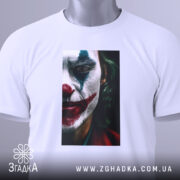 Футболка Джокер білого кольору, складена на поверхні, фон світло-сірий. Принт з обличчям Джокера, видно деталі макіяжу, з етикеткою, що звисає з коміра.