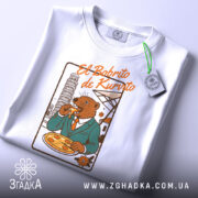 Футболка El Bobrito білого кольору, складена на поверхні, з принтом ведмедя в костюмі, який їсть піцу, з етикеткою на зеленому шнурку.