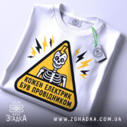 Футболка електрик був провідником. Біла футболка на столі, з принтом у вигляді жовтого трикутника, всередині якого зображений скелет. Фон світло-сірий, а на футболці видно етикетку з зеленим шнурком.