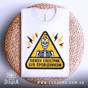 Футболка електрик був провідником. Біла футболка, складена на плетеній основі, з принтом у вигляді жовтого трикутника, всередині якого зображений скелет. Фон світло-сірий.