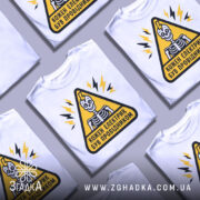 Футболка електрик був провідником. Біла футболка на манекені, з принтом у вигляді жовтого трикутника з чорним контуром, всередині якого зображений скелет з усмішкою. На фоні видно світло-сірий колір. Принт доповнений блискавками.