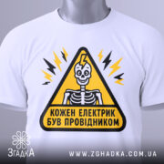 Футболка електрик був провідником. Біла футболка на столі, з принтом у вигляді жовтого трикутника, всередині якого зображений скелет. Фон світло-сірий, а на футболці видно етикетку з зеленим шнурком.