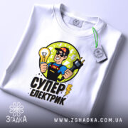 Футболка електрик подарунок, біла, складена на столі, фон світлий, принт з супергероєм, що тримає лампочку та електричний шнур, текст 'СУПЕР ЕЛЕКТРИК' під малюнком, на ярлику зелена стрічка.