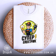Футболка електрик подарунок, біла, складена на плетеній основі, фон світлий, принт з супергероєм, що тримає лампочку та електричний шнур, текст 'СУПЕР ЕЛЕКТРИК' під малюнком.
