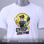 Футболка електрик подарунок, біла, складена на столі, фон світлий, принт з супергероєм, що тримає лампочку та електричний шнур, текст 'СУПЕР ЕЛЕКТРИК' під малюнком, на ярлику зелена стрічка.
