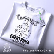 Футболка електрика мистецтво, біла, складена на поверхні, фон світло-сірий, принт чорного кольору з елементами електричних схем і написом 'ЕЛЕКТРИКА ЦЕ МИСТЕЦТВО', на комірці видна етикетка.