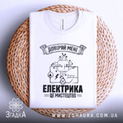 Футболка електрика мистецтво, біла, складена на плетеній кошику, фон білий, принт чорного кольору з елементами електричних схем і написом 'ЕЛЕКТРИКА ЦЕ МИСТЕЦТВО'.