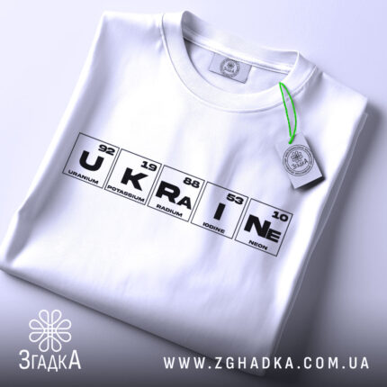 Футболка елементи України, біла, складена на поверхні, фон світло-сірий, принт з елементами таблиці Менделєєва, з зеленим ярликом, що звисає.