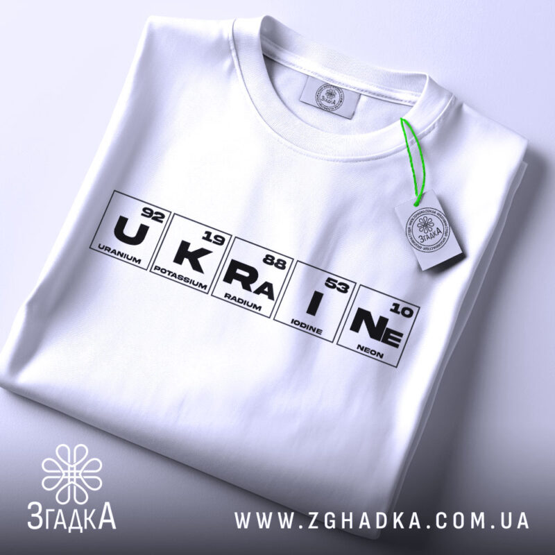 Футболка елементи України, біла, складена на поверхні, фон світло-сірий, принт з елементами таблиці Менделєєва, з зеленим ярликом, що звисає.