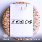 Футболка елементи України, біла, складена на плетеній кошику, фон світло-сірий, принт з елементами таблиці Менделєєва, слово 'UKRAINE' виділено чорним кольором.