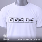 Футболка елементи України, біла, складена на поверхні, фон світло-сірий, принт з елементами таблиці Менделєєва, з зеленим ярликом, що звисає.
