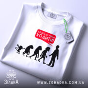 Футболка еволюція досить ходити. Біла футболка, складена на поверхні, фон світло-сірий. Принт з силуетами еволюції та текстом червоного кольору 'Досить за мною ходити!', з зеленим ярликом.