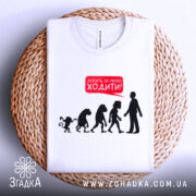 Футболка еволюція досить ходити. Біла футболка, складена на плетеній основі, фон світлий. Принт з силуетами еволюції та текстом червоного кольору 'Досить за мною ходити!'.