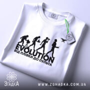 Футболка еволюція, біла, складена на столі, фон світло-сірий, принт чорного кольору з написом 'EVOLUTION ВІД ПЛІВКИ ДО ПУЛЬТА' та силуетами людей, що займаються різними видами зйомки, з етикеткою зеленого кольору.