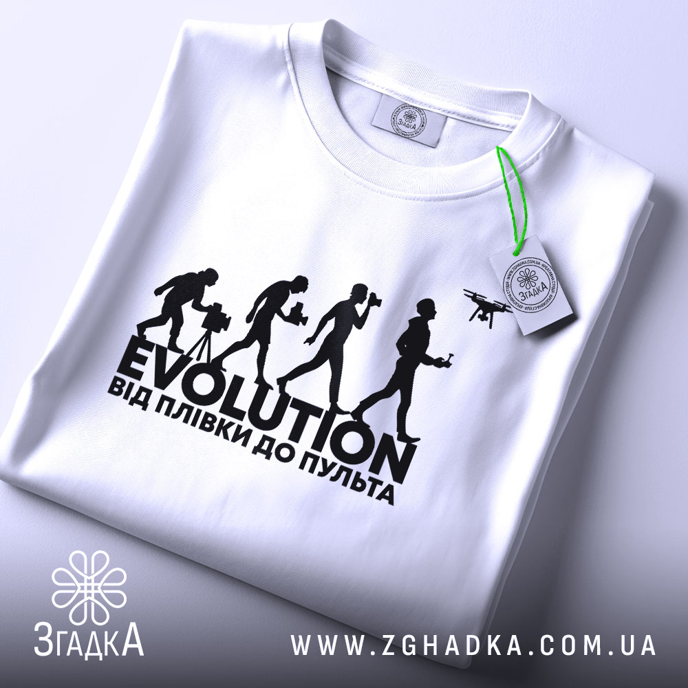 Футболка еволюція, біла, складена на столі, фон світло-сірий, принт чорного кольору з написом 'EVOLUTION ВІД ПЛІВКИ ДО ПУЛЬТА' та силуетами людей, що займаються різними видами зйомки, з етикеткою зеленого кольору.
