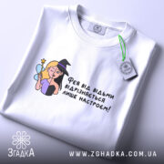 Футболка фея або відьма, білого кольору, зображена зверху, на складеній футболці, фон світлий, принт з ілюстрацією феї та відьми, текст українською мовою, з етикеткою на шнурку.