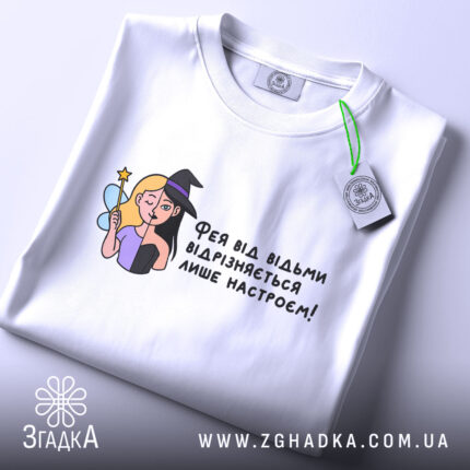 Футболка фея або відьма, білого кольору, зображена зверху, на складеній футболці, фон світлий, принт з ілюстрацією феї та відьми, текст українською мовою, з етикеткою на шнурку.