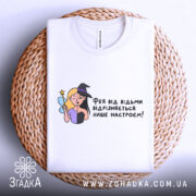 Футболка фея або відьма, білого кольору, зображена зверху на плетеній кошику, фон світлий, принт з ілюстрацією феї та відьми, текст українською мовою.