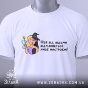 Футболка фея або відьма, білого кольору, зображена зверху, на складеній футболці, фон світлий, принт з ілюстрацією феї та відьми, текст українською мовою, з етикеткою на шнурку.