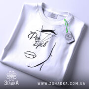 Футболка фея брів, білого кольору, складена на поверхні, фон світлий, принт з малюнком обличчя та написом 'Фея Брів' у чорному кольорі, з зеленим ярликом.