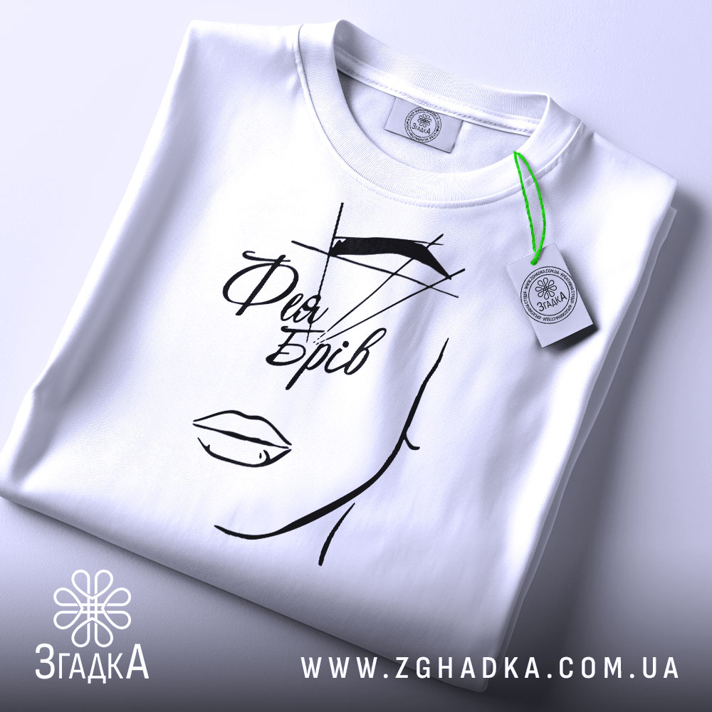 Футболка фея брів, білого кольору, складена на поверхні, фон світлий, принт з малюнком обличчя та написом 'Фея Брів' у чорному кольорі, з зеленим ярликом.